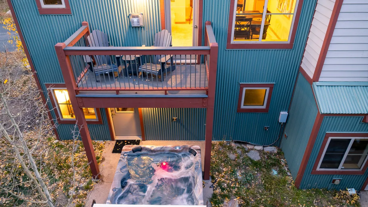 The cozy outdoor balcony features a hot tub with soft ambient lighting, comfortable seating, and large windows revealing the inviting interior warmth. The space is positioned to enjoy views and unwind after a day outdoors.