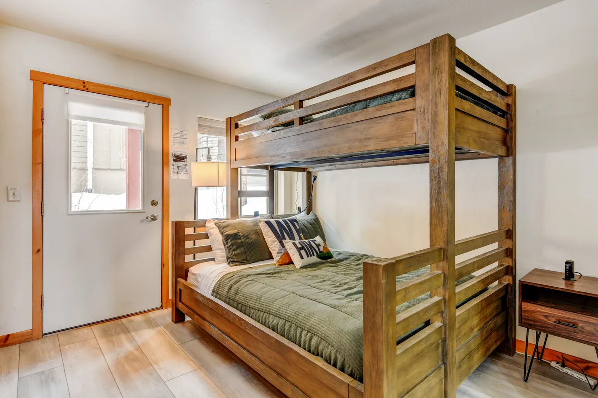 A family-friendly bunk room featuring a twin over queen bunk bed, a window letting in natural light, a lamp, and a convenient nightstand. The room feels inviting for both children and adults, with a playful yet restful vibe.