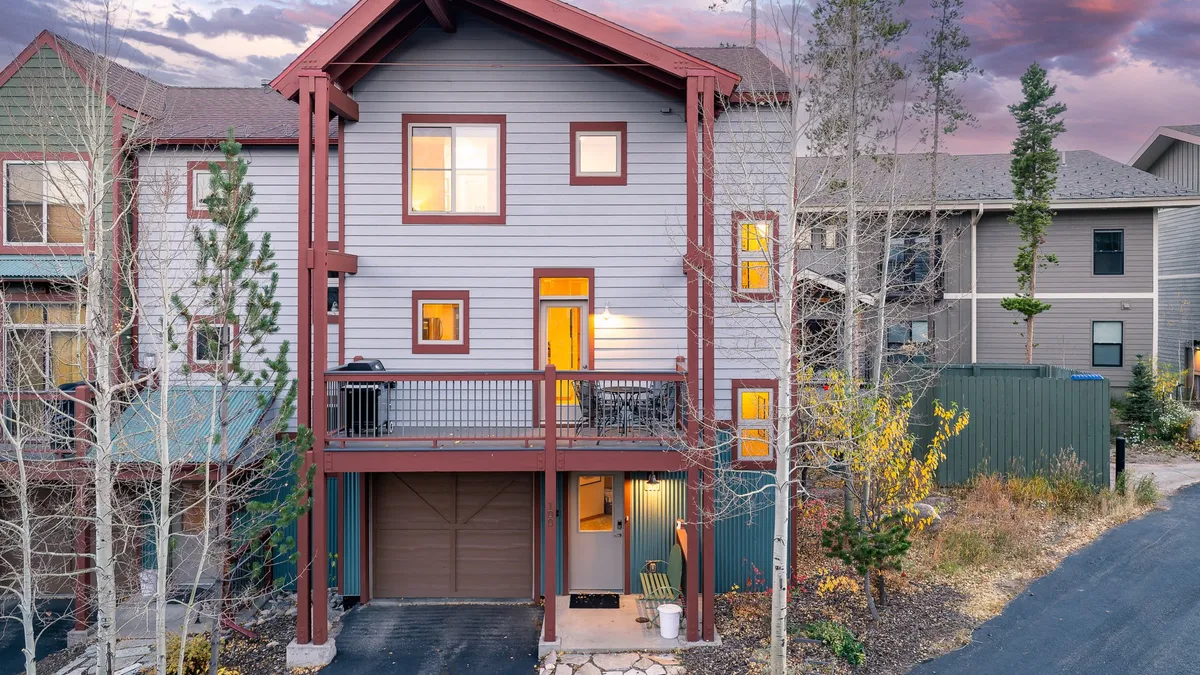 This exterior image displays the chalet’s front porch, a balcony, large windows, and the garage door, set against a welcoming backdrop. The porch and balcony hint at spaces ideal for families to gather before heading out or relaxing after a day of adventure. The proximity to nature is suggested by the landscaping and visible pathways.