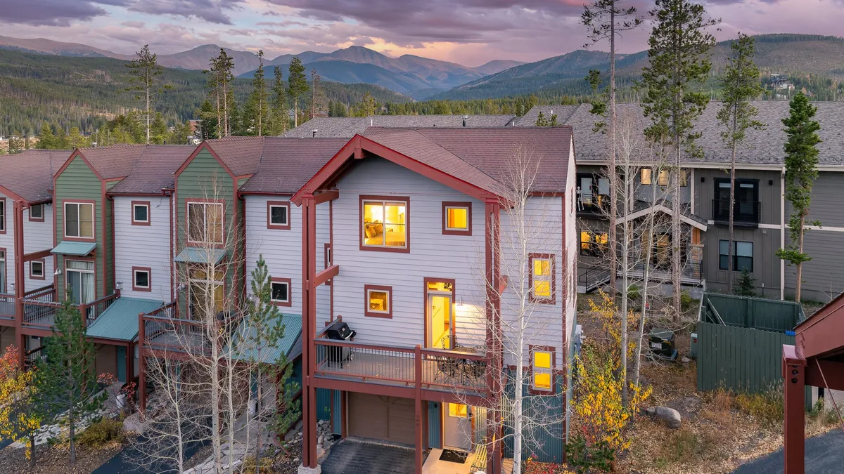 The hero image showcases the inviting front of a luxury chalet nestled among towering trees, with warm lights glowing from the windows, a spacious deck designed for relaxing or gathering, and a backdrop of tranquil mountain scenery perfect for sunsets and stargazing.