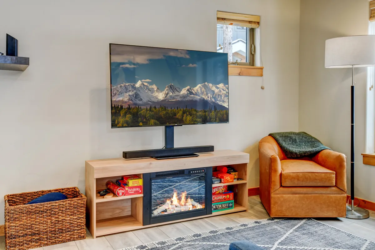 The image showcases a large television displaying a mountain landscape, a comfortable armchair, and a fireplace. The space is inviting and set up for movie nights, board games, and relaxation, encapsulating the multi-functional comfort of the entertainment loft.