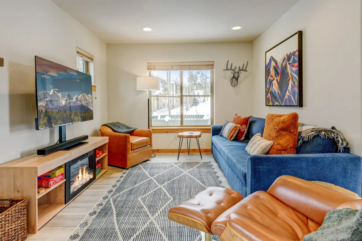 A cozy living room scene featuring a blue couch, a stylish orange armchair, a wooden coffee table, decorative artwork, and a wall-mounted television positioned so that it appears to overlook mountain views. The atmosphere is warm and inviting, perfect for relaxed evenings by the fire or game nights.