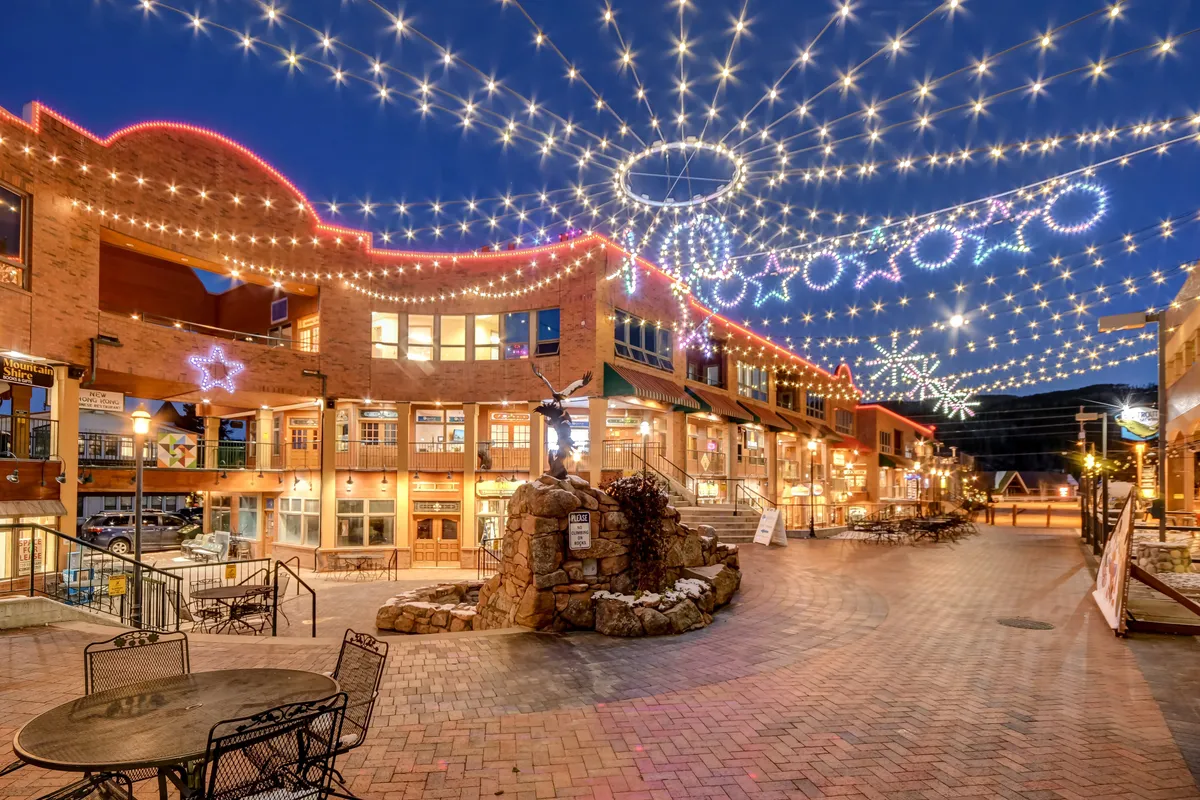 An inviting outdoor plaza scene, featuring string lights overhead, decorative building architecture, an outdoor seating area, and a central fountain or sculpture. The setting is lively and social, hinting at enjoyable evenings and community events.